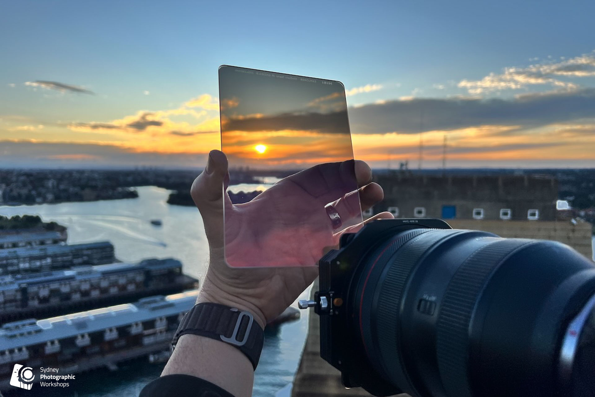 A photographer holding an ND filter up to a Sydney Harbour sunset